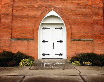 May include: A white double door with black hardware is set in a brick archway. The door is flanked by two bushes and a brick walkway leads to the door.
