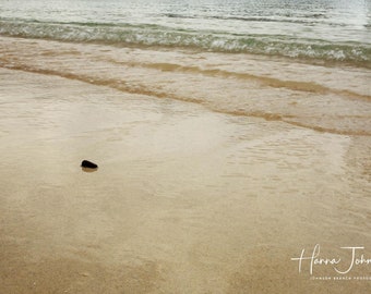 May include: A single dark rock sits on a sandy beach with rippling water in the background. The water is a light brown color and the sand is a light beige color.