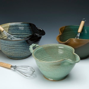 May include: Three ceramic mixing bowls with a unique pour spout design. The bowls are glazed in shades of blue, green, and brown. Each bowl has a wooden handle and comes with a metal whisk.