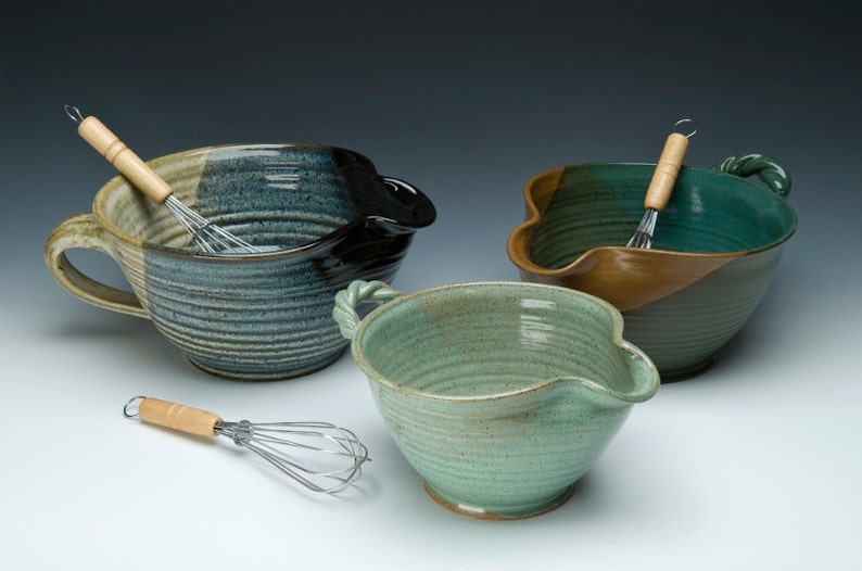 May include: Three ceramic mixing bowls with a unique pour spout design. The bowls are glazed in shades of blue, green, and brown. Each bowl has a wooden handle and comes with a metal whisk.