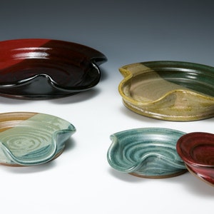 May include: Five ceramic spoon rests in various colors and patterns. The rests are round with a curved indentation for holding a spoon. The colors include red, black, green, brown, and blue.