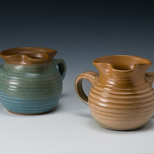 May include: Two ceramic pitchers with brown rims and handles. The pitcher on the left is blue-green with brown stripes. The pitcher on the right is tan with brown stripes.