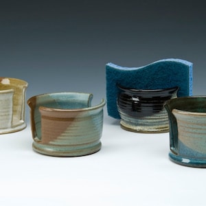 May include: Four ceramic dish soap holders in different colors and glazes. The holders have a slot for a sponge or scrubber. One holder has a blue sponge on top.
