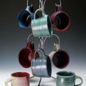 May include: A set of eight ceramic mugs in various colors and patterns, hanging on a metal rack. The mugs are all different shapes and sizes, and some have handles.