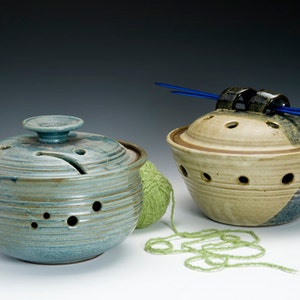May include: Two ceramic yarn bowls with lids. One bowl is blue-green with a lid that has a small knob. The other bowl is tan and brown with a lid that has a metal holder for knitting needles. A ball of green yarn is next to the blue-green bowl.