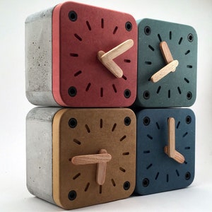 May include: Four square clocks made of concrete and wood. Each clock has a different color face: red, brown, blue, and green. The clock hands are made of wood and are pointing to different times.