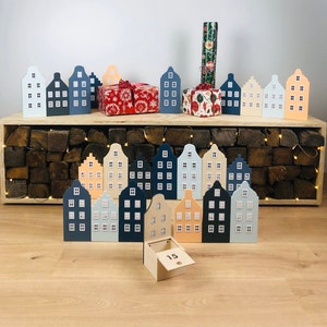 May include: A collection of wooden house-shaped ornaments in various colors, including blue, white, and peach, arranged on a wooden surface. Some houses have small windows. There are wrapped gifts and a wooden box with the number 15.