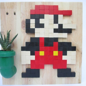 May include: A wooden wall art piece featuring a pixelated image of Super Mario, made with small wooden blocks painted red, black, and white. A green plastic pipe with a small succulent plant is attached to the left side of the wall art.