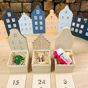 May include: Wooden advent calendar featuring miniature house-shaped boxes in various pastel colors. Each box has a hinged lid and contains a small toy or ornament. Numbered wooden tiles are placed in front of the boxes.