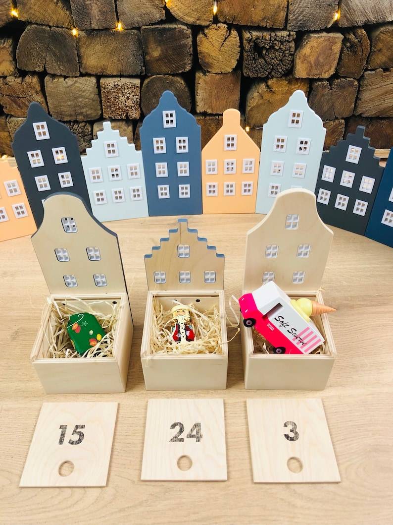May include: Wooden advent calendar featuring miniature house-shaped boxes in various pastel colors. Each box has a hinged lid and contains a small toy or ornament. Numbered wooden tiles are placed in front of the boxes.