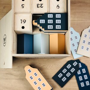 May include: A wooden advent calendar with numbered drawers and house-shaped ornaments. The calendar has a natural wood finish, with black numbers on the drawers. The house ornaments are in various colors, including navy, light blue, and peach.