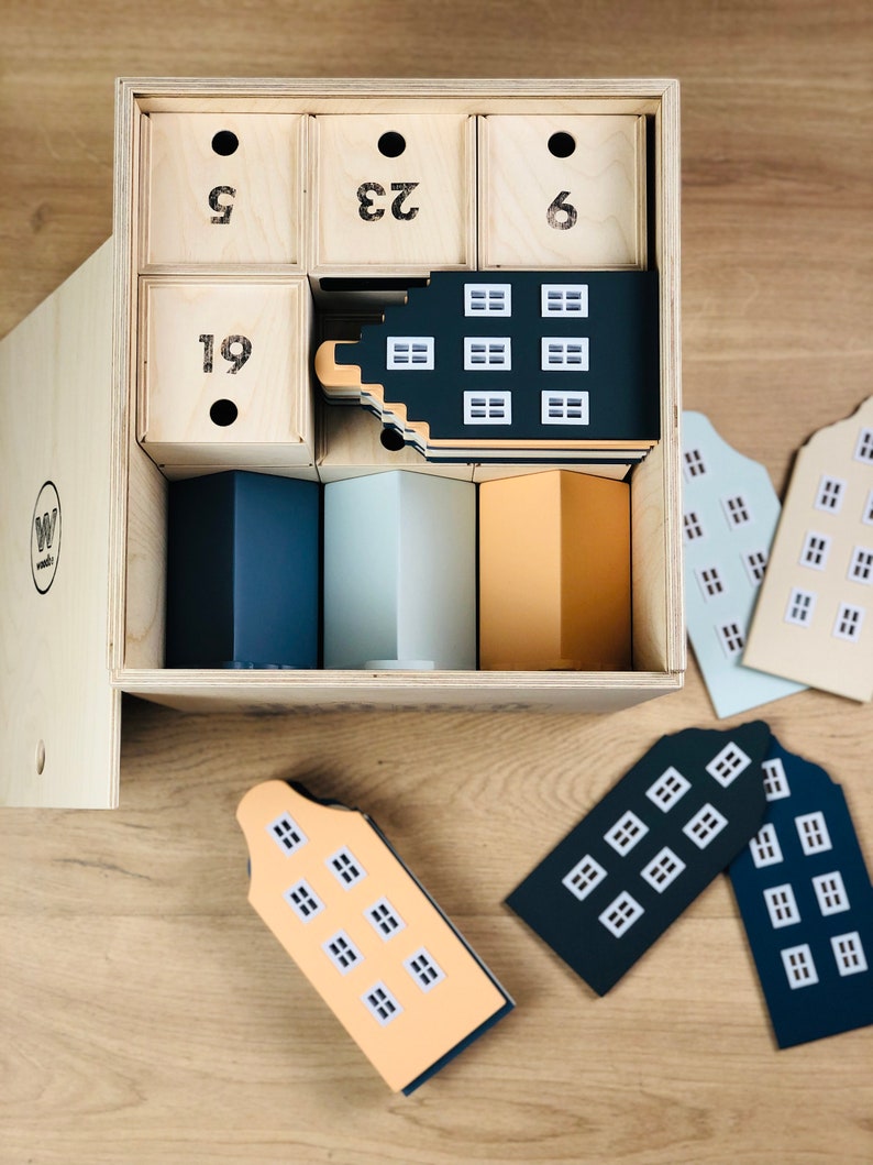May include: A wooden advent calendar with numbered drawers and house-shaped ornaments. The calendar has a natural wood finish, with black numbers on the drawers. The house ornaments are in various colors, including navy, light blue, and peach.
