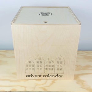 May include: A light-colored wooden advent calendar box with a lid. The front of the box features an etched design of four stylized houses and the words "advent calendar" below. The lid has a small hole and a logo.