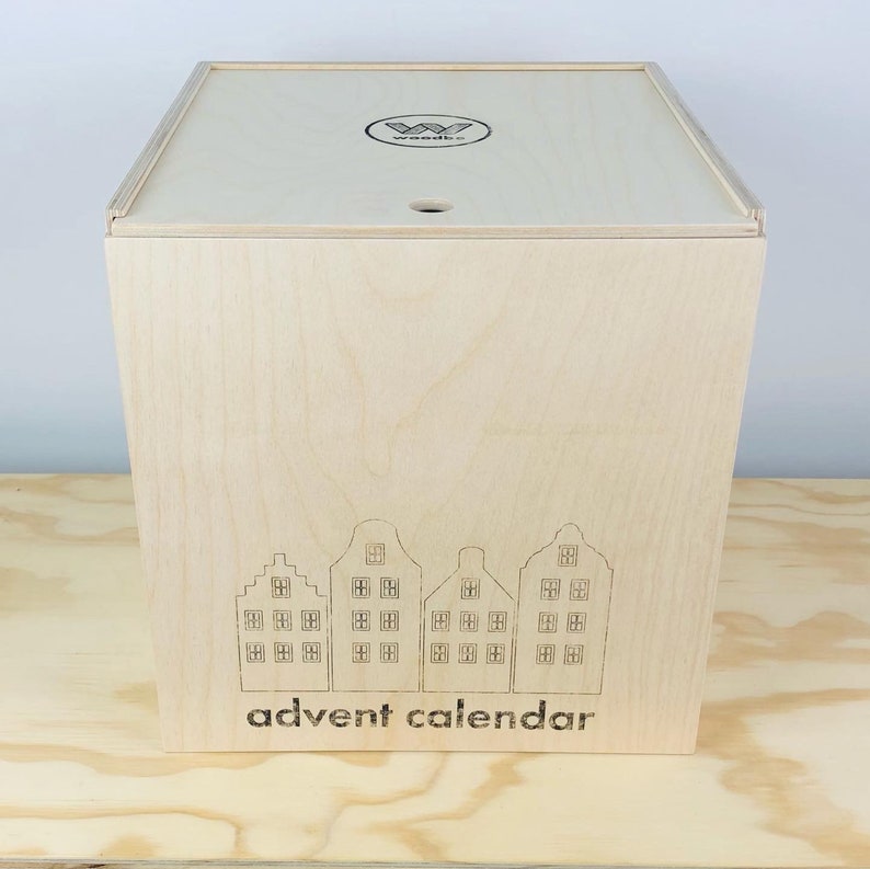 May include: A light-colored wooden advent calendar box with a lid. The front of the box features an etched design of four stylized houses and the words "advent calendar" below. The lid has a small hole and a logo.