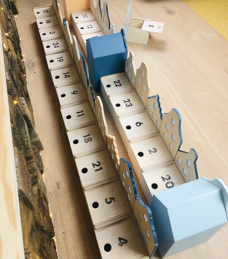 May include: Wooden advent calendar with numbered boxes in a town design. The boxes are arranged in rows, with numbers printed on them. The calendar has a light blue and beige color scheme, with a few blue houses.