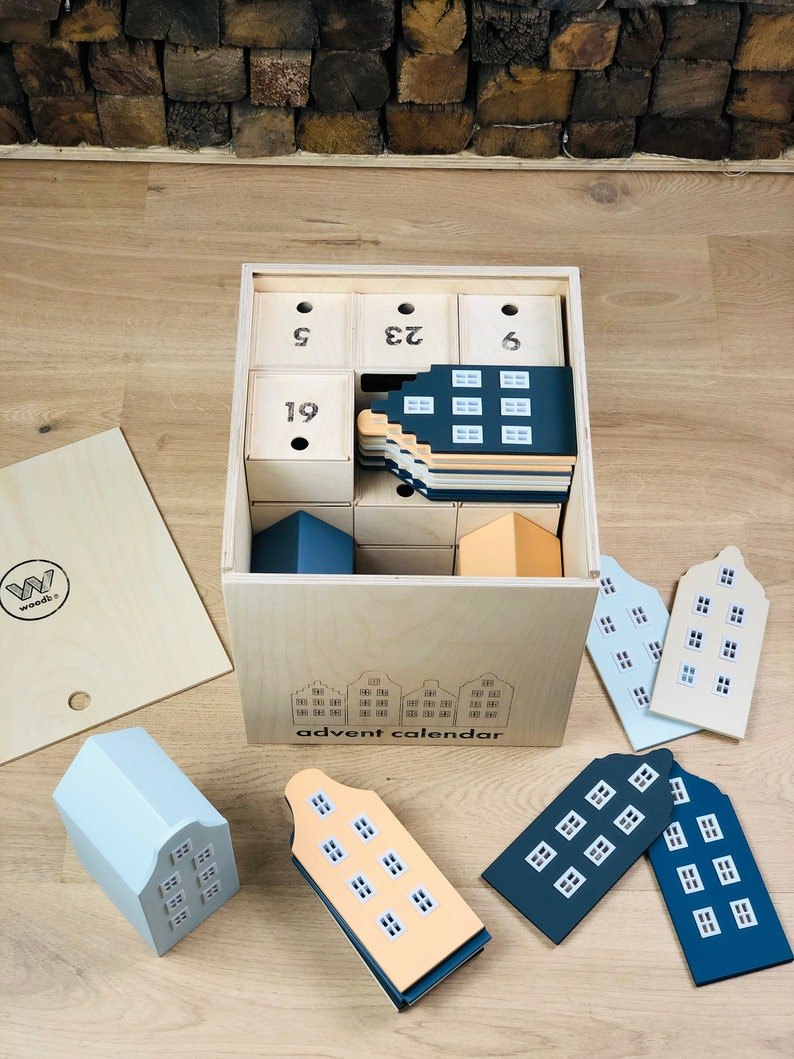 May include: Wooden advent calendar with a light wood finish. The calendar features a box with small drawers and house-shaped ornaments in various colors, including blue, orange, and white. The box is labeled "advent calendar".