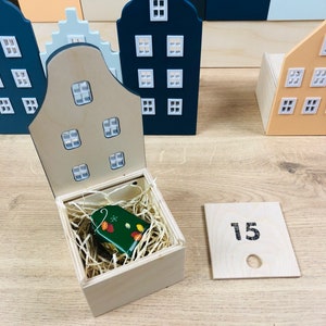 May include: Wooden miniature house figurines in various colors, including blue, beige, and white, with white window details. A small wooden box contains a green tag with leaf designs. A wooden number 15 sign is also present.
