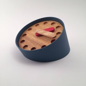 Op de afbeelding: Een blauwe en houten klok met een rode en houten uurwijzer. De klok heeft een uniek ontwerp met een schuin gezicht.