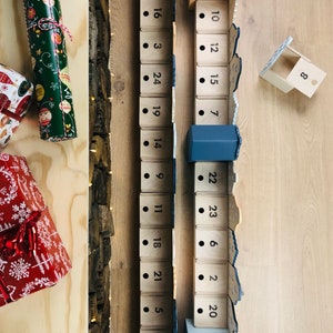 May include: Wooden advent calendar with numbered boxes, some open. The calendar is in a wooden frame with a rustic, textured side. Wrapped gifts in green and red paper are on the left.