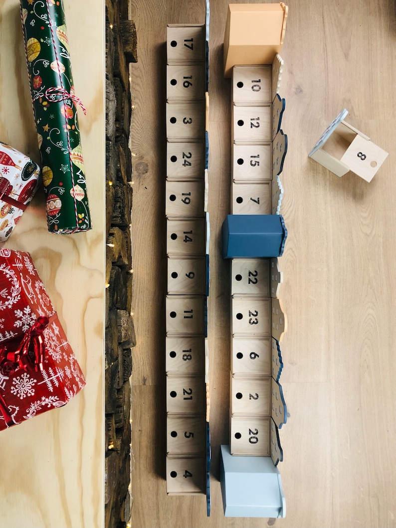 May include: Wooden advent calendar with numbered boxes, some open. The calendar is in a wooden frame with a rustic, textured side. Wrapped gifts in green and red paper are on the left.