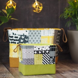 Puede incluir: Dos cestas de almacenamiento de tela con diseños de patchwork. La cesta más grande es amarilla, gris y blanca. La cesta más pequeña es verde, negra y blanca.