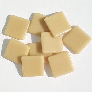 May include: A collection of ten square, light beige glass tiles. Each tile has rounded corners and a glossy finish. The tiles are scattered on a white surface, with shadows adding depth. Ideal for mosaic projects or decorative crafts.