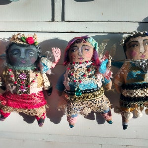 May include: A collection of handcrafted fabric dolls, each with a painted face and intricate, vibrant attire. The dolls feature lace, beads, and crowns, showcasing a whimsical, artistic style. They are arranged against a white backdrop, highlighting their unique designs.