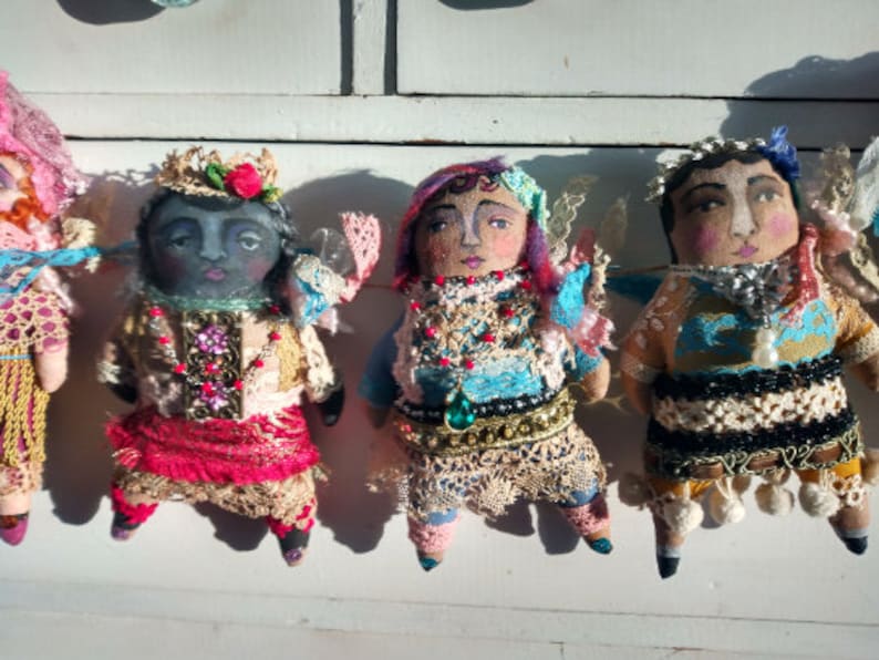 May include: A collection of handcrafted fabric dolls, each with a painted face and intricate, vibrant attire. The dolls feature lace, beads, and crowns, showcasing a whimsical, artistic style. They are arranged against a white backdrop, highlighting their unique designs.