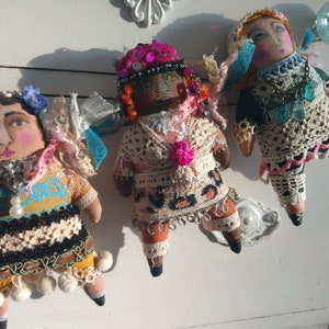 May include: Three handcrafted fabric dolls with painted faces and elaborate clothing. The dolls feature unique outfits with crochet, lace, and beaded details. Each doll has a distinctive hat or head covering, set against a white backdrop.