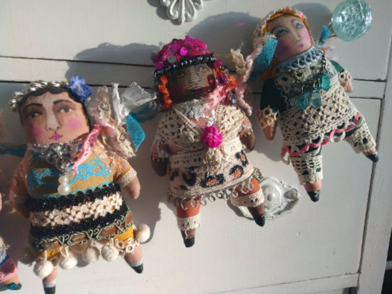 May include: Three handcrafted fabric dolls with painted faces and elaborate clothing. The dolls feature unique outfits with crochet, lace, and beaded details. Each doll has a distinctive hat or head covering, set against a white backdrop.