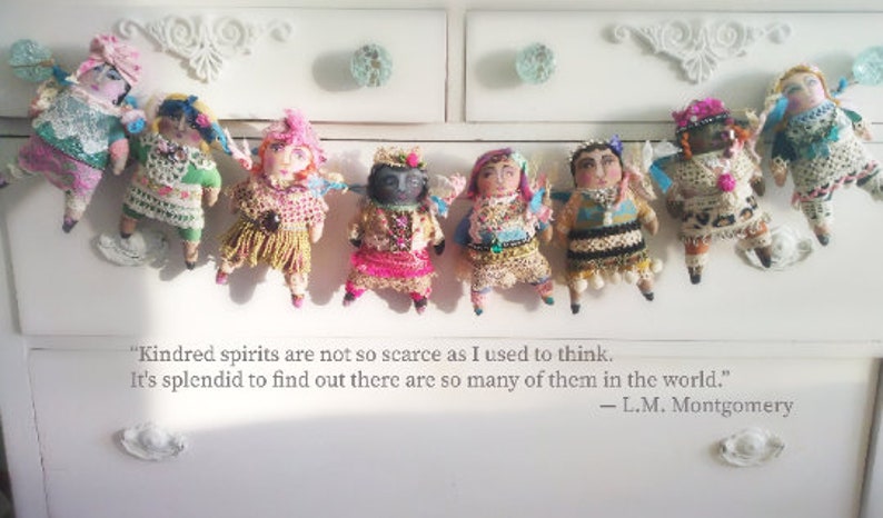 May include: Handmade dolls with wings, each with a unique outfit of colourful fabrics, beads, and lace. They are displayed on a white chest of drawers, with a quote by L.M. Montgomery.