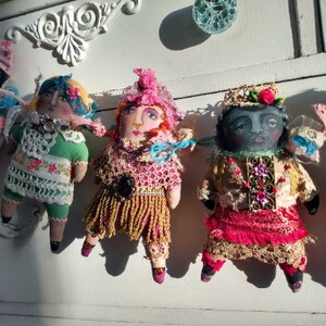 May include: A collection of handmade fabric dolls with painted faces and detailed clothing. Each doll has a unique design, featuring lace, embroidery, and colourful fabrics. The dolls are arranged in a line, showcasing their individual styles and craftsmanship.