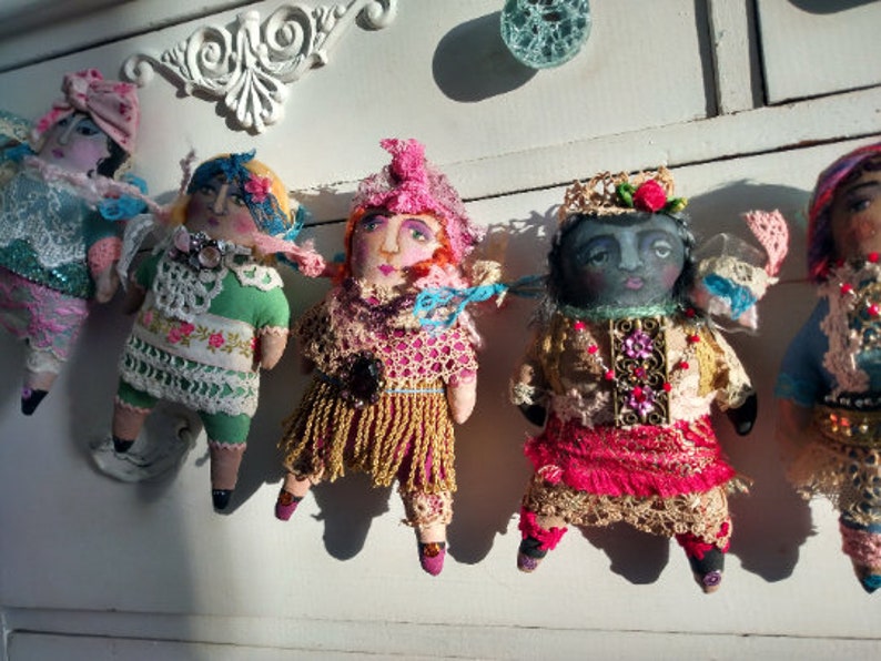 May include: A collection of handmade fabric dolls with painted faces and detailed clothing. Each doll has a unique design, featuring lace, embroidery, and colourful fabrics. The dolls are arranged in a line, showcasing their individual styles and craftsmanship.