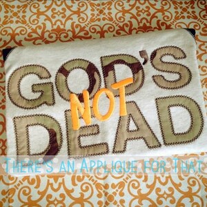Puede incluir: Tela color crema con las palabras "GOD'S NOT DEAD" aplicadas en tela de camuflaje y naranja. La frase "THERE'S AN APPLIQUE FOR THAT" est&aacute; escrita debajo del texto principal.