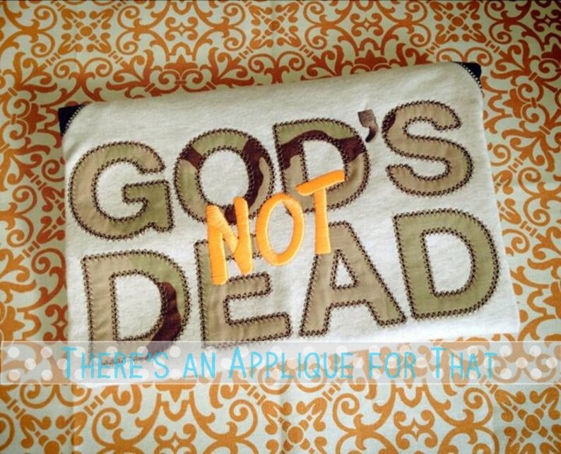 Puede incluir: Tela color crema con las palabras "GOD'S NOT DEAD" aplicadas en tela de camuflaje y naranja. La frase "THERE'S AN APPLIQUE FOR THAT" est&aacute; escrita debajo del texto principal.