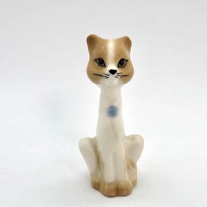May include: A ceramic cat figurine with a tan and white color scheme. The cat has a long neck, a blue dot on its chest, and is sitting upright. The cat's face has black whiskers and eyes.