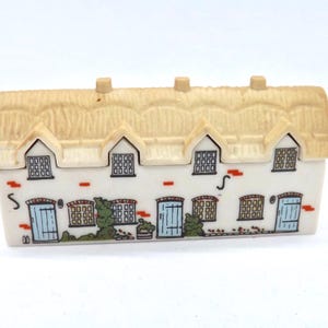 Puede incluir: Figura de cerámica de una casa de campo con tejado de paja. La fachada blanca presenta puertas azules, ventanas y detalles de ladrillo. Arbustos verdes y un pequeño jardín pintados en la base.