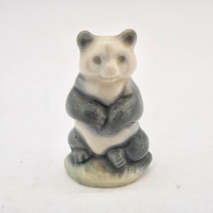 May include: A small ceramic panda bear figurine. The panda is depicted in a seated position with its arms crossed. The bear has a white face and body with grey markings. It is sitting on a small, green base.