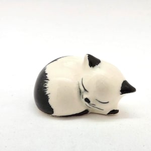 May include: A ceramic figurine of a sleeping cat. The cat is curled up, with its eyes closed. The cat is white with black markings on its ears and back. The figurine is on a white surface.