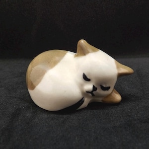 May include: A ceramic figurine of a sleeping cat. The cat is curled up, with its eyes closed. The cat is white with brown markings on its head and ears. The figurine is on a dark surface.