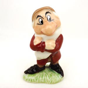 May include: A ceramic figurine of a character with a large nose, thick eyebrows, and a tan cap, wearing a dark red tunic with a white collar and black shoes. The character stands with arms crossed over the chest on a green painted base.