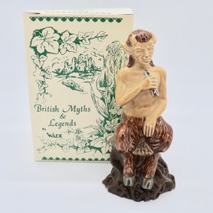 May include: A ceramic figurine of a brown and tan faun playing a flute. The figurine is sitting on a brown base and is part of a collection called "British Myths & Legends by Wade".