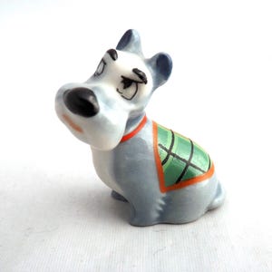 May include: A ceramic dog figurine in a seated position, featuring a light blue body, dark blue ears, and a black nose. It wears a green and orange plaid blanket and a red collar. The figurine has a vintage, cartoon style.