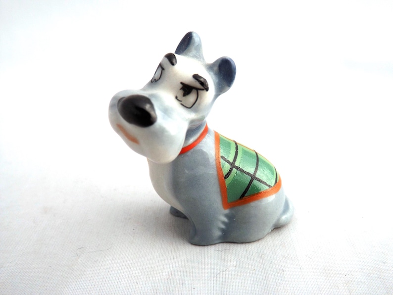 May include: A ceramic dog figurine in a seated position, featuring a light blue body, dark blue ears, and a black nose. It wears a green and orange plaid blanket and a red collar. The figurine has a vintage, cartoon style.