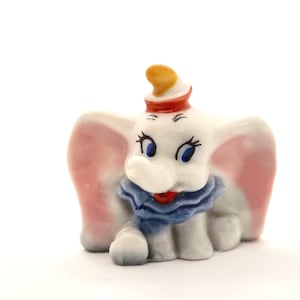 May include: A small ceramic figurine of a baby elephant with large pink ears, a blue ruffled collar, and a red and yellow hat. The elephant has blue eyes, long black eyelashes, and a grey body, depicted in a seated position with a trunk.