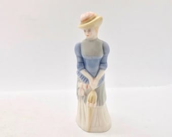 Może przedstawiać: Porcelanowa figurka kobiety w długiej sukni i kapeluszu, trzymającej parasol. Figurka jest pomalowana pastelowymi kolorami, z niebieskim stanikiem, szarymi rękawami i kremową spódnicą. Kapelusz jest żółty z różowym akcentem.