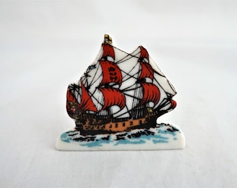 Könnte beinhalten: Eine weiße Keramikfigur eines Schiffes mit roten Segeln und einem braunen Rumpf. Das Schiff segelt auf blauem Wasser mit weißen Wellen.