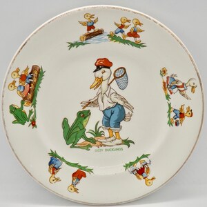 Può includere: Un piatto in ceramica bianca con un'illustrazione colorata di anatre e rane. Il piatto presenta un'anatra con un berretto rosso e una rete, e il testo "Dizzy Ducklings".