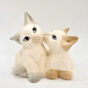 May include: A ceramic figurine of two stylized cats. One cat is light gray and white, sitting upright. The other cat is light tan and white, kissing the first cat. The cats have black eyes and are on a white background.
