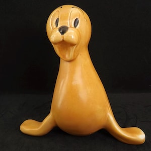 Peut inclure: Figurine de phoque en céramique de couleur brun doré. Le phoque a une expression souriante, avec des yeux noirs et un nez noir. Ses nageoires sont étendues et la figurine est en position assise. Le fond est sombre.